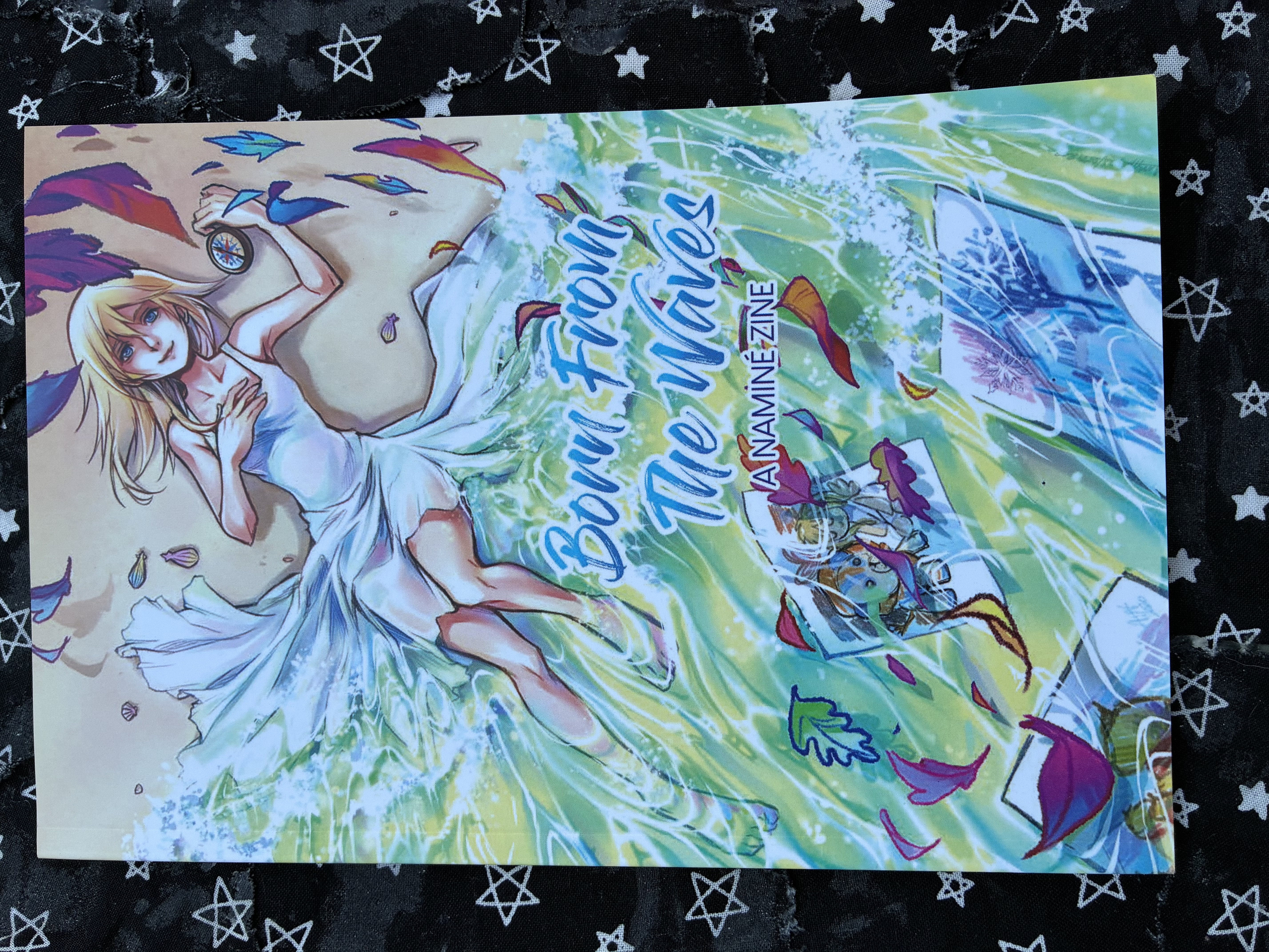 Front of Born from the waves: a namine zine with Namine laying half in the sand and waves surrounded by her drawings and holding a compass.